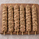 Six raw XL Mediterranean chicken kebabs with a dry green herb seasoning, arranged vertically on a rustic wooden board.